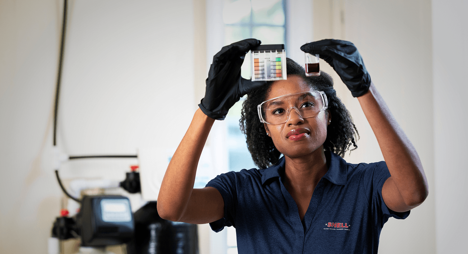A Snell technician in a blue uniform and safety gear compares a water sample to a testing chart in front of a water softener system.