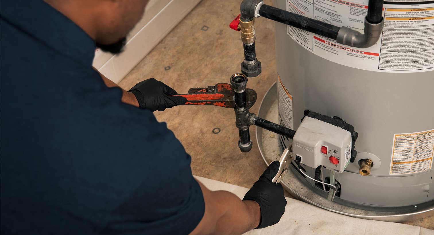 A Snell plumber wearing black gloves uses a pipe wrench to repair the gas line and control valve assembly on a residential water heater tank.